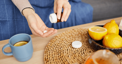 Manos de mujer sujetando vitaminas para el cansancio sobre una mesa con frutas e infusiones